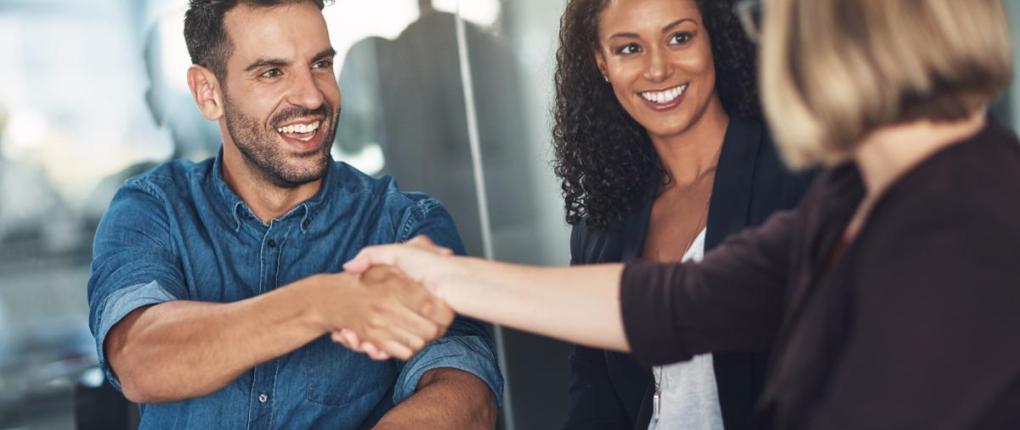 Three professionals sit together in a meeting, smiling as two people shake hands across a table in an office setting.
