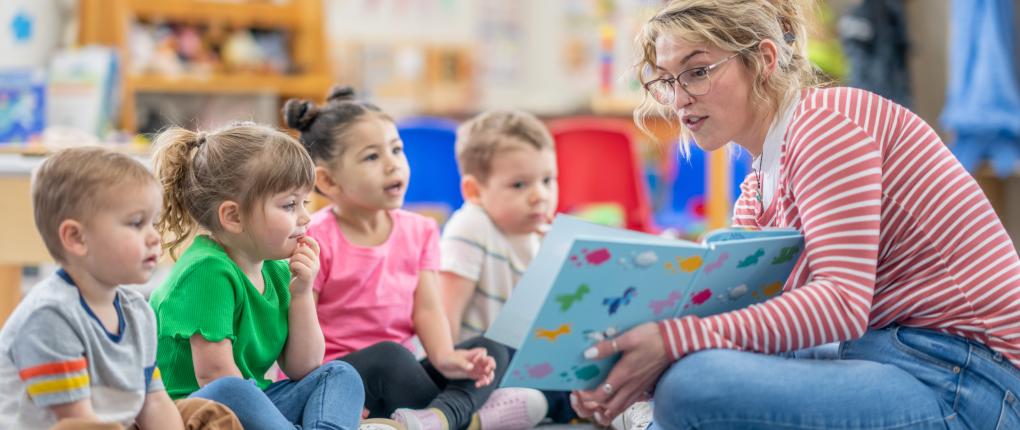 A preschool teacher sits on the floor of her classroom with a small group of students as she reads them a book. The children are each dressed casually and are focused on the story.