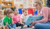 A preschool teacher sits on the floor of her classroom with a small group of students as she reads them a book. The children are each dressed casually and are focused on the story.