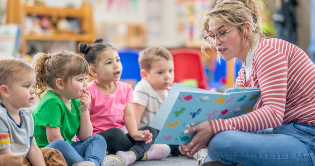 A preschool teacher sits on the floor of her classroom with a small group of students as she reads them a book. The children are each dressed casually and are focused on the story.