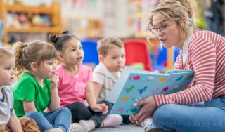 A preschool teacher sits on the floor of her classroom with a small group of students as she reads them a book. The children are each dressed casually and are focused on the story.